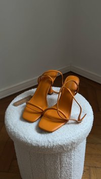 Sandalias de tacón cuadrado naranja con tiras finas expuestas sobre un taburete redondo blanco texturizado, contra un suelo de madera y una pared gris.