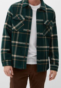 Uomo che indossa una giacca a quadri verde scuro e beige con bottoni sopra una camicia bianca, con pantaloni marroni, in piedi con le braccia rilassate.