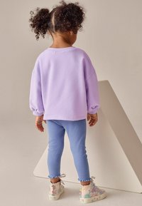 Hanorac supradimensionat lavandă, colanți luminoși albaștri cu margine destrămată și adidași cu model și accente pastelate. Copil văzut din spate.