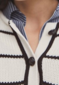 Cardigan blanc en maille avec des rayures noires, doté de grands boutons noirs. Associé à une chemise col à rayures bleues et blanches en dessous.
