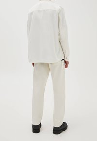 Jasny, oversize koszula i pasujące spodnie z miękkiego materiału, w zestawie z czarnym obuwiem. Ubranie charakteryzuje się prostym fasonem i brakiem widocznych wzorów.