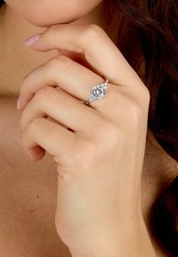 Anello con diamante a forma di pera incastonato in argento, con piccole pietre di accento ai lati, esposto su una mano con smalto neutro.