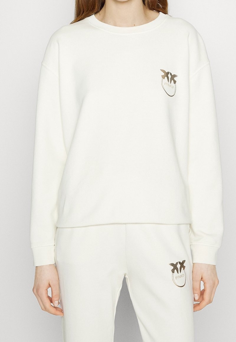 Woman wearing a white sweatshirt and pants with matching small gold bird logos on the chest and thigh.