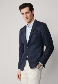 Námořnický blazer s pruhovaným vzorem v texturované tkanině, s jedním knoflíkem a náprsní kapsou. Nositý přes světle modrou polokošili a krémové kalhoty.