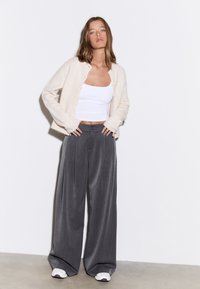 Cardigan crème, débardeur blanc ajusté, pantalon gris à jambes larges avec plis, et baskets blanches. Textures lisses et design minimaliste.