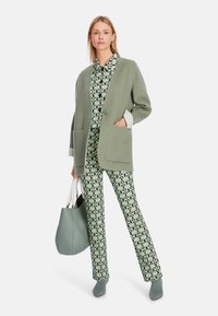 Grüner übergroßer Blazer über einer gemusterten Bluse und passenden Hosen, kombiniert mit einer großen grauen Handtasche und Stiefeletten.