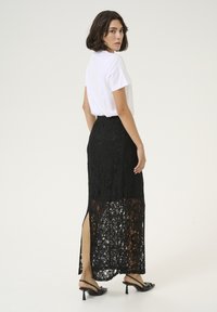 Jupe maxi en dentelle noire avec une fente latérale, ornée d'un motif floral texturé. Assortie à un t-shirt blanc et des sandales à talons noires.