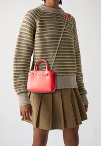 Person wearing a striped beige and yellow sweater, olive pleated skirt, and red Guess handbag with chain strap across the torso.