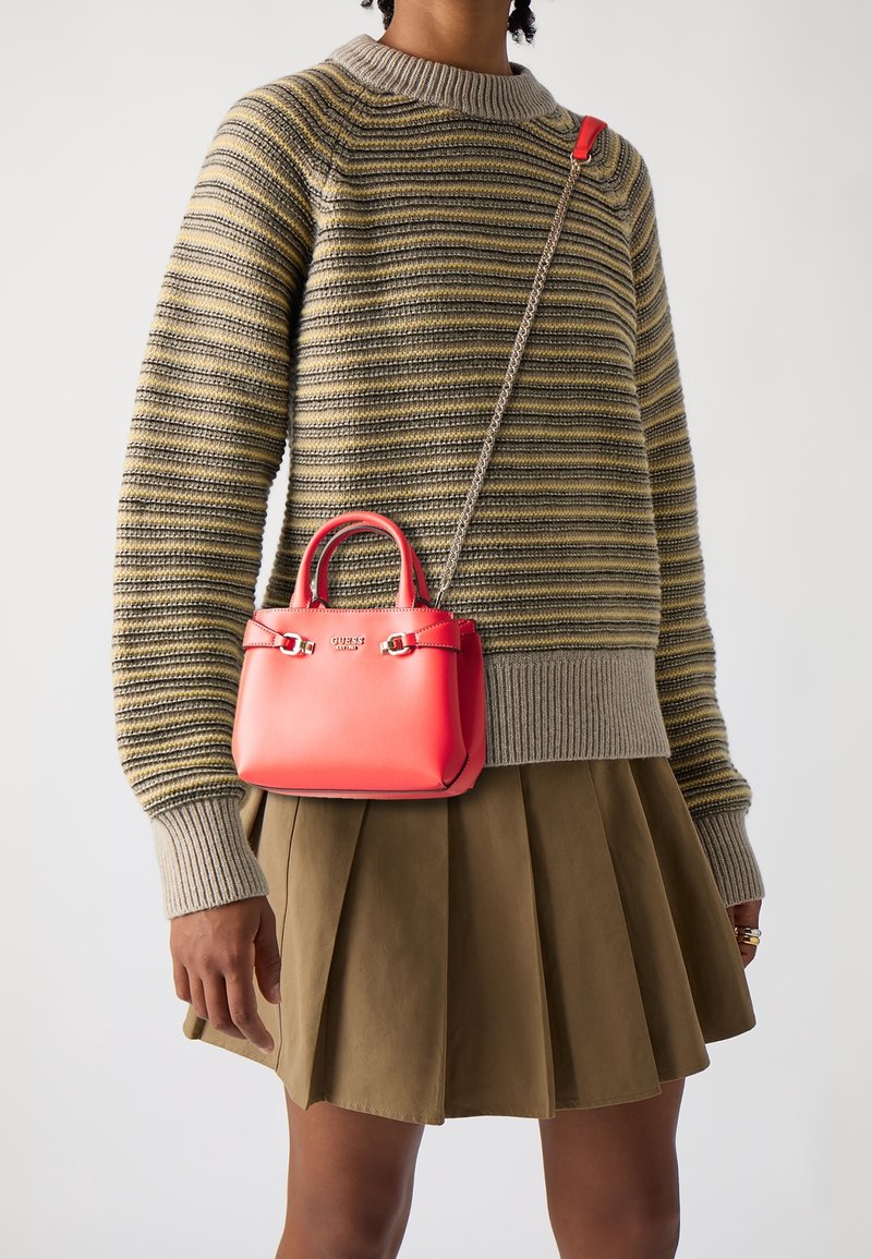 Person wearing a striped beige and yellow sweater, olive pleated skirt, and red Guess handbag with chain strap across the torso.