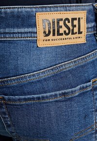 Jeans de mezclilla azul con un parche de cuero etiquetado como "DIESEL" en la parte trasera, con bolsillos cosidos y un diseño clásico de cinco bolsillos.