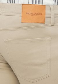 Pantalon en coton beige avec une poche arrière, comportant une étiquette en cuir marron embossée du texte « JACK&JONES » et des détails de couture.