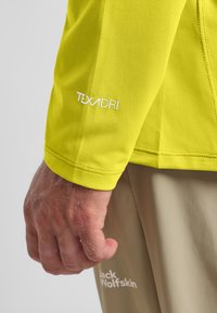 Geel long-sleeve top met een gestructureerde stof, een strakke manchet, en een witte "TEXADRI" logo op de mouw, gecombineerd met lichtbeige broek.