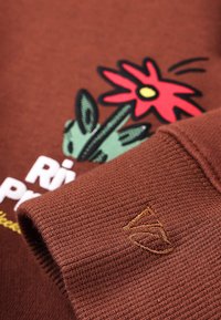 Smeđa majica dugih rukava s cvjetnim uzorkom koji prikazuje crveni cvijet i zelene lišće. Rukavi s rebrastim manžetama i izvezenim detaljem logotipa. Tekst u bijeloj i žutoj boji.