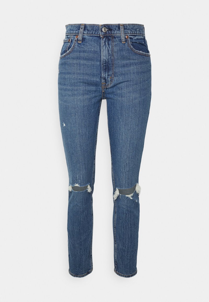 Jeans de ganga azuis com perna reta, cintura alta, rasgos nos joelhos desgastados e costura em contraste. Apresenta cinco bolsos e fecho de botão metálico.