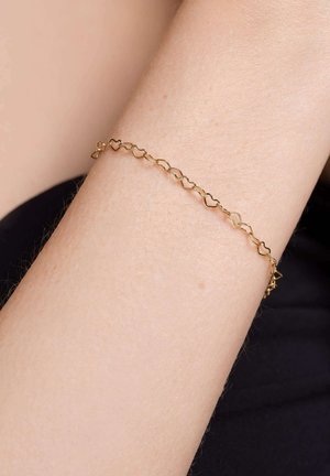 Gouden armband met aan elkaar geschakelde hartvormen. Gladde textuur met een delicate uitstraling, rustend op lichte huid tegen een donkere achtergrond.