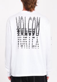 Hvid langærmet T-shirt lavet af bomuld, med et dristigt sort grafisk design på ryggen, der indeholder abstrakt tekst og former.