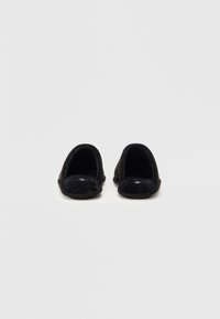 Zapatillas de lana negras con forma redondeada, textura suave y diseño de abertura trasera. Cuenta con un tejido texturizado y una suela de goma negra.