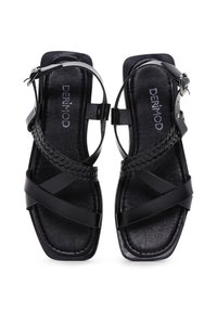Sandalias negras con detalle trenzado, punta cuadrada, suela plana y correa en el tobillo. Hechas de piel suave con un acabado mate. Nombre de la marca visible en el interior.