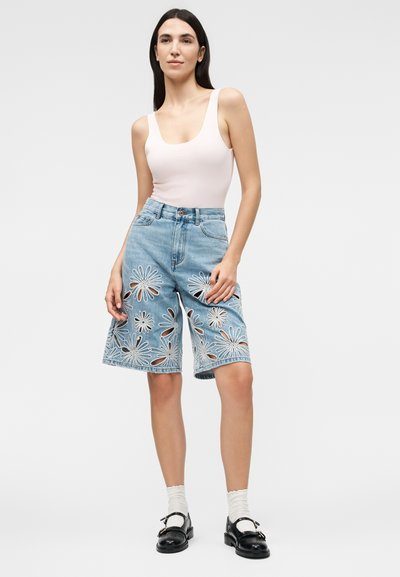 Γυναίκα που φοράει λευκό αμάνικο τοπ και τζιν σορτς μέχρι το γόνατο με μεγάλα φλοράλ κοψίματα, λευκές κάλτσες και μαύρα loafers.