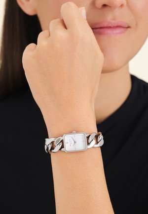 La montre argentée présente un cadran blanc rectangulaire avec des marqueurs horaire minimalistes, monté sur un bracelet en maillons métalliques chunky.