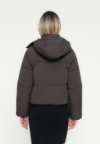 Veste matelassée noire courte avec capuche, présentant un design matelassé, des manches longues et une coupe courte, affichée sur un fond blanc.