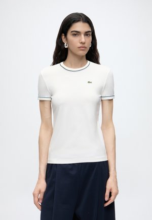 Femme aux longs cheveux foncés portant une chemise blanche à manches courtes avec des bordures foncées et un pantalon bleu marine, debout devant un fond clair uni.
