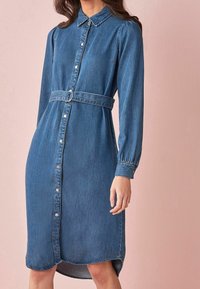 Robe-chemise en denim bleu moyen, avec un devant boutonné, des manches longues, un col et une taille ceinturée avec une grosse boucle. L'ourlet est légèrement arrondi.