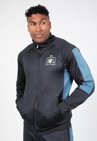 Zwarte zip-up trainingsjas met blauwe accenten, met een logo op de borst, zijzakken en een hoge kraag. Soepele, atletische stof.
