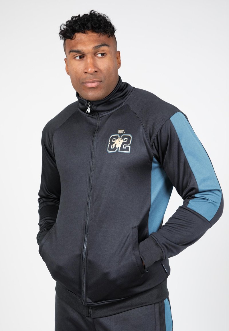Zwarte zip-up trainingsjas met blauwe accenten, met een logo op de borst, zijzakken en een hoge kraag. Soepele, atletische stof.