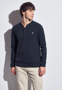 Navy long-sleeve henley shirt with three buttons and a textured finish. Features a small logo patch on the chest. Paired with light beige pants.