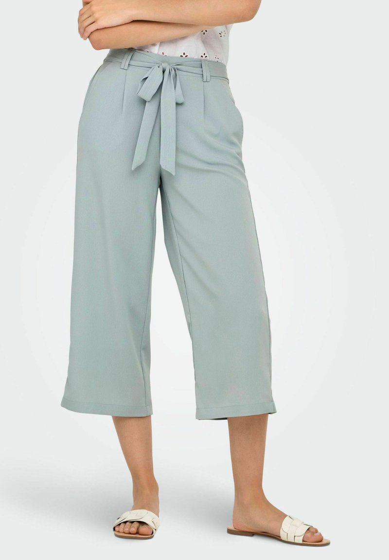 Pantalon large couleur bleu clair en tissu lisse. Comprend une taille ceinturée avec un nœud et des revers retroussés. Associé à des sandales blanches à enfiler.