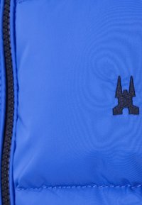 Blå puffjacka med en slät textur, svart dragkedja och en liten svart logotyp som liknar en silhuett av ett slott.