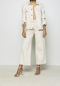 Kerman denim takki napilla, joka on yhdistetty vastaaviin leveälahkeisiin housuihin. Mallilla on jalassaan läpinäkyvillä hihnoilla varustetut korkokengät. Pehmeä, teksturoitu kangas.