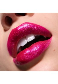 Jasno różowe, błyszczące usta z brokatową teksturą, prezentujące pełny kształt i gładką powierzchnię. Częściowo widoczne białe zęby w tle.