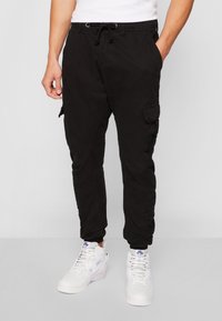 Zwarte cargobroek met elastische tailleband en trekkoord, voorzien van steekzakken aan de zijkant en een losse pasvorm, gecombineerd met witte hoge sneakers.