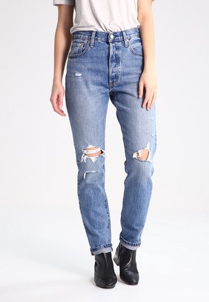 Kobieta ubrana w jasnoniebieskie przetarte dżinsy z podartymi kolanami, białą włożoną do środka koszulkę i czarne botki na kostkę na jednolitym tle.