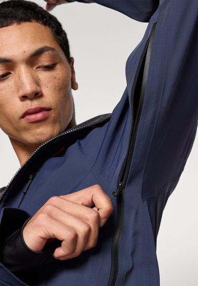 Marineblaue wasserdichte Jacke mit glatter Oberfläche, ausgestattet mit einem Reißverschluss-Unterarmventil und schwarzen Reißverschlussakzenten. Nahaufnahme einer Hand, die den Reißverschluss greift.