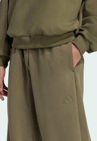 Conjunto deportivo verde oliva que incluye un sudadera y pantalones a juego, fabricado en una tela suave con un dobladillo acanalado y un acento del logo de Adidas.