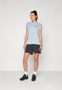 Valkoinen, logoilla varustettu, ilman hihoja oleva vaaleansininen t-paita, yhdistettynä tummiin shortseihin, joissa on graafinen kuvio. Mallilla on päällään mustat tenniskenkä ja valkoiset nilkkasukkahousut.