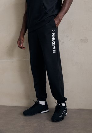 Sorte joggerbukser med hvid "PUMA CREW '48" tekst på venstre side, parret med sorte sportssko og hvide sokker. Glat stoftekstur.