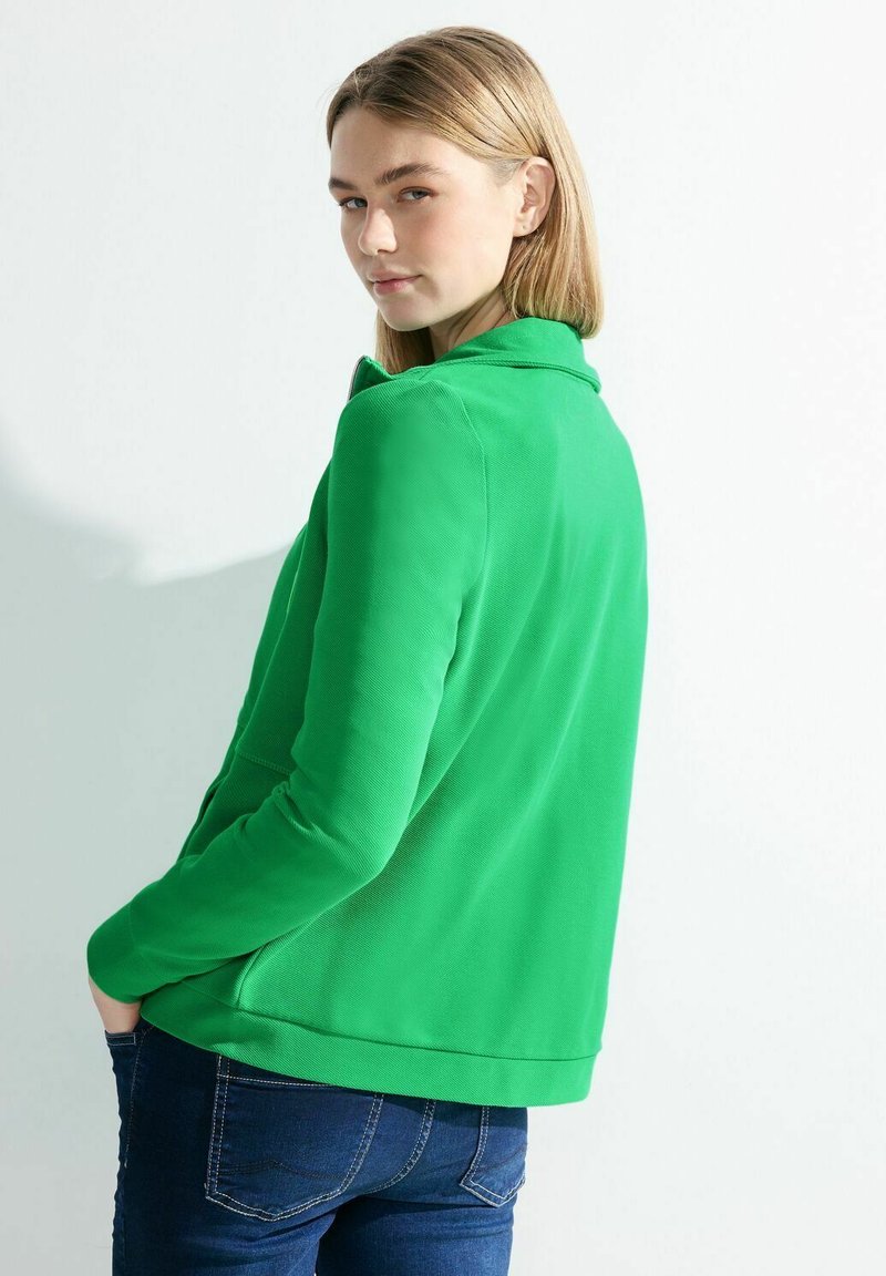 Giacca verde in tessuto morbido con colletto, tasche frontali e texture liscia, abbinata a jeans blu scuri. Vista laterale del modello.