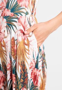 Combinaison fleurie en tissu blanc texturé avec des motifs floraux roses et bleus. Comprend une poche latérale et une coupe décontractée.