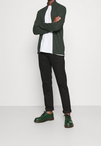 Mörkgrön zip-up kofta över en vit t-shirt, i kombination med svarta byxor och gröna läderskor. Enkla design och åtsittande silhuett.