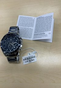 Orologio cronografo con quadrante texturizzato nero, cassa e bracciale in acciaio inossidabile argentato, etichetta di garanzia attaccata e manuale utente aperto accanto ad esso.