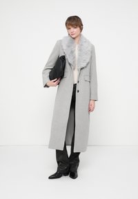 Abrigo largo gris con un collar de piel sintética, dos bolsillos frontales y un corte recto. Modelo sosteniendo un bolso negro, vistiendo pantalones oscuros y botas.