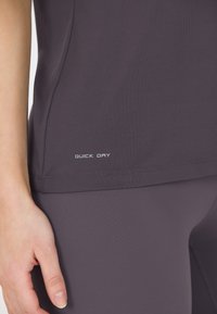 Tumman violetti nopeasti kuivuva urheilu paita, jossa on sileä rakenne ja logo helman kohdalla. Lähikuva paidasta ja käsi lepäämät lähettyvillä.