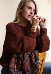 Blonde vrouw in een bruine trui zit op een houten stoel, steunt met haar armen op de leuning, met een bruin-blauwe geruite sjaal eroverheen gedrapeerd.