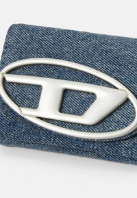 Denim peňaženka so strieborným kovovým logom v oválnom tvare a ostrými detailmi, s textúrovaným modrým vzorom látky.