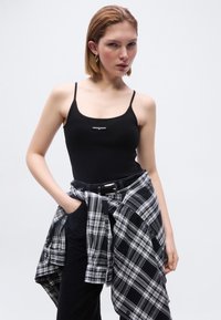 Femme portant un débardeur noir à fines bretelles avec le logo "tommy jeans", un pantalon noir, et une chemise à carreaux noir et blanc nouée autour de sa taille.