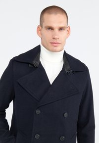 Marineblauwe, dubbelzijdige wollen peacoat met grote zwarte knopen en een leren accentkraag, gedragen over een witte coltrui.